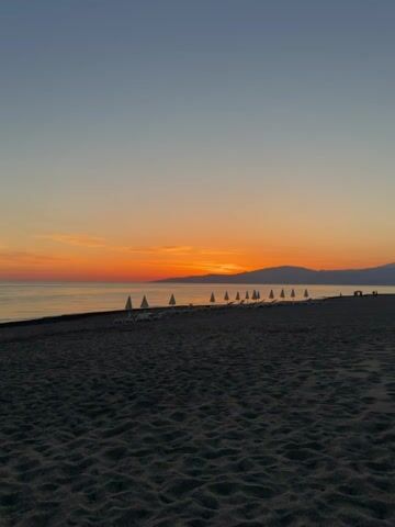 The video shows a wide shot of a beach at sunset. The sand is dark, with visible footprints. A line of closed white beach umbrellas and lounge chairs stands parallel to the shoreline. The sea is calm, reflecting the orange and yellow hues of the setting sun. A silhouette of distant mountains is visible on the horizon, underneath the vibrant sunset. The sky transitions from a light blue to a deeper orange near the horizon.
