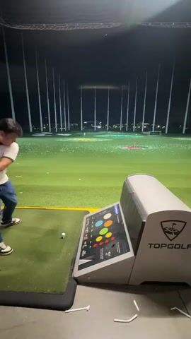 A man in a white t-shirt and jeans is playing golf at Topgolf. He is swinging a golf club, and the video shows the golf ball flying through the air. The scene is at night, and the golf course is lit up with lights. There are other golf balls scattered on the course, and there are also some targets in the distance. The man is standing on a green mat, and there is a console with buttons in front of him.