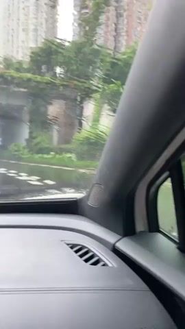 The video shows a view from inside a car driving on a wet road in what appears to be a city. The road is marked with white lines, and there are puddles of water on the surface. Several cars are visible on the road, including a white SUV that is ahead of the camera car. Buildings and trees can be seen on either side of the road, and the sky is overcast. It is raining, and water is running down the windshield. The camera appears to be mounted on the dashboard. The overall impression is one of a rainy day drive through an urban environment.