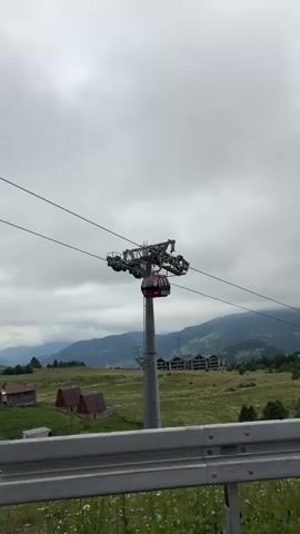 The video shows a cable car moving along its cable against a cloudy sky. Below, there are rolling green hills dotted with small houses and some trees. In the foreground, a metal guardrail is visible. The camera perspective suggests it is moving, possibly from inside a vehicle. The scene transitions to a view of a winding road through a forested area, with a road sign indicating a curve ahead. The sky remains overcast throughout.