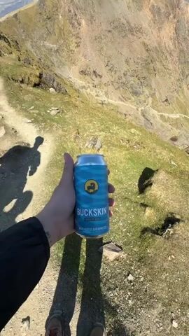The video shows a person holding a can of Buckskin lager on top of a mountain. The landscape is rocky and grassy, with a clear blue sky above. The person is standing on a path and the shadow of the person is visible. There are other mountains in the distance.