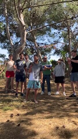 A group of seven young men of varying heights, builds, and attire stand in a clearing covered in pine needles, with several tall trees and some scattered vegetation in the background. The man in the center, wearing a gray and white shirt and patterned swim trunks, holds what appears to be a small plant or flower in his hand and smiles. The other men, dressed in shorts, t-shirts, and casual wear, are gesturing and looking at the camera. A few of them are pointing or striking poses, suggesting a lighthearted and playful mood. The lighting indicates it is a sunny day.  A decorative net with ribbons hangs between two trees in the background.