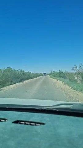 The video shows a view from inside a vehicle, looking forward through the windshield. A road stretches ahead into the distance, bordered by sparse vegetation on either side under a clear blue sky. A white truck is visible on the road, approaching the viewer's vehicle. The dashboard of the vehicle is visible in the foreground, in shades of teal and gray. The video captures the truck as it gets closer.