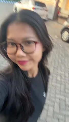 A young woman with long, dark hair and glasses is filming herself in an outdoor setting. She is wearing a black t-shirt with a white logo on the left chest. The background shows a paved area with brick-like stones, and a vehicle with a red and white graphic is visible in the upper right portion of the frame. Another vehicle appears to be parked in the distance. The lighting suggests it is daytime.