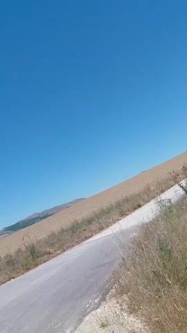 The video shows a rural landscape with a bright blue sky and a winding road. A sign with a red circle and two black cars inside is visible on the side of the road. A green van drives away from the camera down the road. Fields of dry grass and some green vegetation are visible on either side of the road, with rolling hills and mountains in the distance. The sun is shining, creating a clear and bright atmosphere.