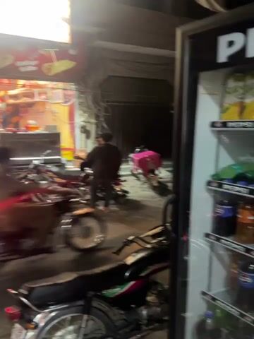The video captures a lively street food establishment at night, likely in Model Town, Pakistan. The scene is dominated by a brightly lit shop with a prominent sign reading "بسم اللہ ولی برگر پوائنٹ" (Bismillah Wali Burger Point) in Urdu, along with English slogans like "Best Burger in the Town" and "HOME DELIVERY." Several phone numbers (0306-4418937, 0303-0044911) are also displayed. The shop's facade features images of various food items such as burgers, shawarmas, and biryani.

Outside the shop, a row of motorcycles is parked, suggesting a common mode of transport for customers or delivery riders. There's a noticeable presence of people gathered around the entrance and inside the shop, appearing to order or wait for food. The atmosphere is bustling, indicative of a popular late-night spot. At the beginning of the video, a person on a motorcycle with a pink delivery bag passes by, reinforcing the impression of active food delivery services. The ground is paved, and the area is illuminated by the shop's lighting and streetlights, creating a vibrant nocturnal setting.