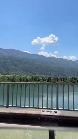The video shows a car driving across a bridge on a clear, sunny day. The water below the bridge is a vibrant turquoise color. In the background, there are large, green mountains. The camera angle shifts from the side of the bridge to the interior of the car, showing the driver's side mirror and then the road ahead. Other cars are visible on the highway.