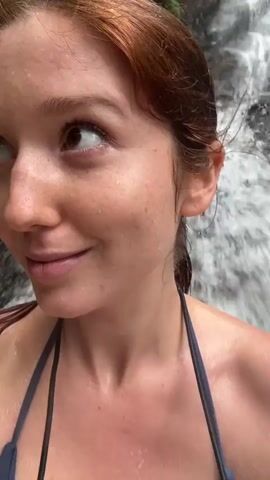 A woman with reddish-brown hair and freckles stands in front of a waterfall, smiling slightly at the camera. She is wearing a blue swimsuit. The waterfall is behind her, with white water cascading down. The scene appears to be taken outdoors in a natural setting.