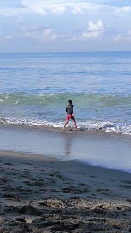 The video shows a person walking into the ocean. The person is wearing what appears to be a long-sleeved black top and red shorts, and the ocean's water is shallow enough that they are able to walk a fair distance before diving in. The water is a clear blue-green color. There are small waves breaking. The beach has dark sand, and there are shadows cast on the sand. In the background, the sky is blue with some white clouds.