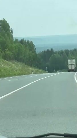 The video shows a view from inside a car driving on a highway. The road has two lanes in the direction of travel, separated by a solid white line. On the left side of the road, there is a grassy embankment covered with trees. In the distance, there are more trees and rolling hills. A white truck with a trailer is visible in the right lane ahead, and two cars are driving in the left lane ahead of the truck. The sky is overcast.