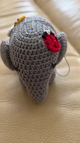 A gray crocheted ghost-shaped object with small, crocheted embellishments is lying on a beige leather surface. The object has a ladybug and a bee attached to it. A thin gray thread is visible extending from the object. The lighting is soft, and the focus is on the object.