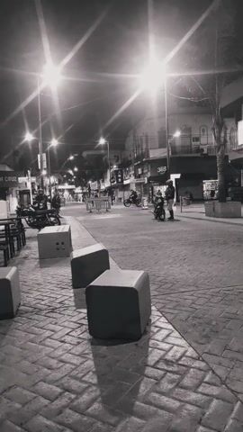The video presents a nighttime scene of a street in black and white. Streetlights are prominent, casting bright rays. The ground is paved with patterned bricks. There are cube-shaped concrete blocks along the side of the street. Several parked motorcycles are visible near the street's edge. In the distance, there are buildings with signs and lights. A person is walking on the sidewalk.