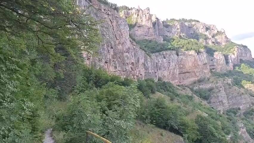 The video shows a drone shot panning across a rocky cliff face and a lush green valley. The cliff is tall and steep, with visible layers of rock and vegetation growing on its ledges. Below the cliff, a river winds through the valley, and a small village with red-tiled roofs is nestled amongst the trees. The sky is partly cloudy, with patches of blue visible. The overall impression is one of natural beauty and a peaceful rural setting.