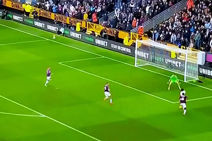 The video shows a soccer game in progress. The field is green and surrounded by a crowd of spectators. A soccer ball is in play near the goal. There are several players on the field, some dressed in white and others in maroon and white. The goalkeeper, wearing a green shirt, is down on the ground attempting to block a shot. The goal is behind him with "UNIBET" displayed prominently on the net. Signage reading "DEWALT" and "UNIBET" lines the sidelines. One of the players in white kicks the ball towards the goal. The other players are running and positioned around the goal.