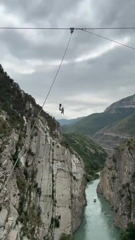 This video shows a zipline ride over a river canyon. A person is attached to a zipline cable, suspended over the river. The person appears to be enjoying the ride, as they are smiling and waving. The canyon walls are steep and rocky, with lush green vegetation growing on the slopes. The river below is a bright turquoise color, and there is a small raft floating on the water. The sky is cloudy and overcast, but the light is still bright enough to see the details of the landscape.