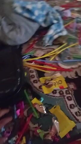 The camera pans up from a partially open dark grey suitcase filled with small plastic objects on a rug to a chaotic scene on the rug. The rug has a patterned design. There are scattered drinking straws of various colors (yellow, red, green, blue), small plastic toy pieces (some resemble shoes), yellow snack packages, a container of strawberry yogurt, small paper cards with lettering, two black smartphones, and a white power adapter connected to one phone. A section of a light blue blanket is visible in the upper left. A hand with pale skin and a manicured nail is briefly seen near the suitcase at the beginning of the video. The general impression is of a messy space.