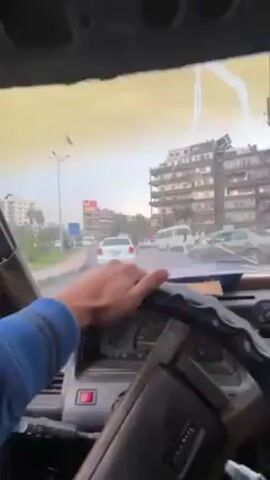 The video, shot from inside a vehicle, shows a person driving on a city street. The driver's left hand is visible on the steering wheel. The dashboard of the vehicle is old and worn. The windshield is dirty. Outside, there are cars and buildings, typical of an urban landscape. The sky is overcast.