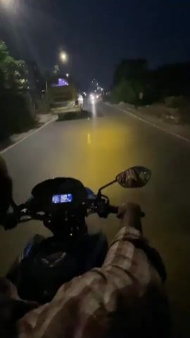 A person is riding a motorcycle at night. Their hands are on the handlebars, and they are wearing a brown and white checkered shirt. The motorcycle is blue and black, and the dashboard shows a speed of 47. In front of them, a yellow bus is visible on the left side of the road, and an ambulance with flashing lights approaches on the right side. Other vehicle lights are also visible in the distance. Streetlights illuminate the dark road.