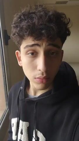 A young man with curly dark hair and light skin is recording a close-up video of himself. He is wearing a black hooded sweatshirt and appears to be standing indoors in a hallway or room with neutral-colored walls. He touches his hair once.  A window is visible to his left, and a chair is visible behind him in the hallway. The lighting is soft.