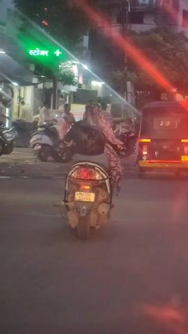 A video shows a motorcycle traveling down a street. The motorcycle is being ridden by two people. The person in front is wearing a pink and white patterned headscarf and the person in the back is wearing a black jacket and dark pants. The motorcycle is beige and has a license plate that reads "MH10 DN 6764". Behind the motorcycle are a few other vehicles, including a tuk-tuk and a silver SUV. The street is paved and there are buildings and trees lining the sides. The lighting suggests it is dusk or early evening.