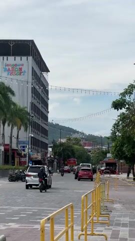 The video shows a street scene with buildings and cars. On the left side, a tall building with the sign "RISON RESORT" is visible, along with palm trees. A white SUV and a person on a motorcycle are driving on the road. In the center, there are more cars and trees. On the right side, a person wearing a hat and a bright vest is sweeping the sidewalk. Several concrete blocks are placed along the sidewalk. The sky is cloudy. The video pans slightly to the right, revealing more of the buildings and the street. Advertisement billboards are attached to the buildings on the right.
