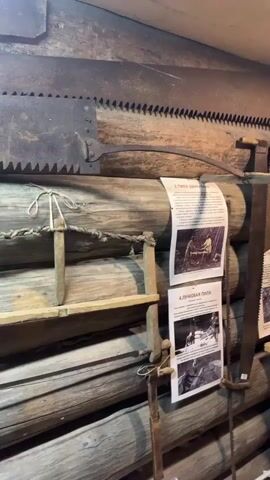 The video pans across a display of antique woodworking tools mounted on a rustic wooden wall. Large logs are stacked behind the tools. The tools include various saws, hand planes, and other implements, all appearing to be made of wood and metal. Some of the tools are displayed on a wooden workbench in the foreground, where papers with text and images are also placed. The environment suggests a museum exhibit or a historical workshop, with rough-hewn wooden walls and beams. The lighting is adequate, with a visible light fixture overhead. There are no people visible in the video.