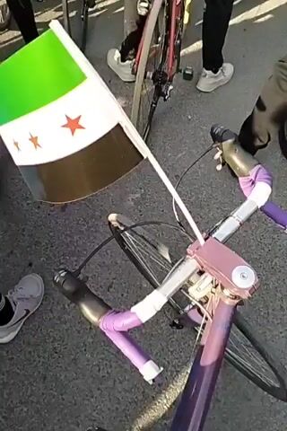 A crowd of cyclists wearing helmets gathers beneath an inflatable archway, seemingly preparing to embark on a ride. Many are astride their bicycles.  One bicycle, featuring a light purple frame, is adorned with a Syrian flag attached to its handlebars.  The environment appears to be an urban street, with buildings visible in the distance beyond the archway. The day seems bright and clear.