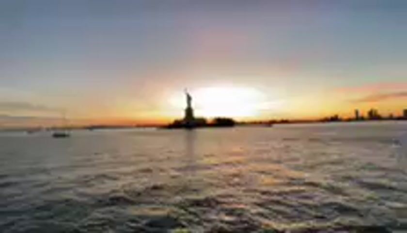This media file captures a sunset view of the Statue of Liberty from Jersey City, United States. The iconic statue is silhouetted against a vibrant sky painted with hues of orange, pink, and yellow, indicative of either sunrise or sunset. The sun itself is a bright, almost blinding orb positioned just behind the statue, casting a warm glow across the water.

The scene is set on a body of water, likely the Hudson River or New York Harbor, with gentle waves rippling across the surface. In the distance, a city skyline, presumably Manhattan, is visible as a dark silhouette against the colorful sky. To the left of the Statue of Liberty, a sailboat or two can be seen, adding a touch of tranquility to the expansive view.

The overall mood is serene and majestic, evoking a sense of awe inspired by the grandeur of the landmark and the beauty of the natural light. The time of day is clearly either dawn or dusk, with the soft, warm light bathing the scene. The weather appears to be clear, allowing for an unobstructed view of the horizon. There is no visible text in this media file.