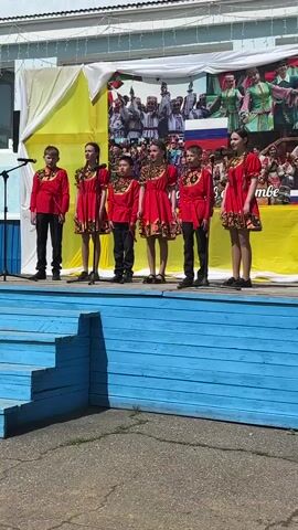 Six children, three boys and three girls, stand on a blue wooden stage in front of a backdrop banner. They are dressed in matching red outfits with black pants for the boys and short red dresses for the girls. The outfits feature a black and gold design at the neckline and waist. They are singing into microphones. Behind them, the backdrop displays a photo collage of people in traditional clothing, flags, and text in a language that appears to be Russian. The stage is set outdoors on a sunny day. The scene suggests a performance or celebration of culture or heritage.