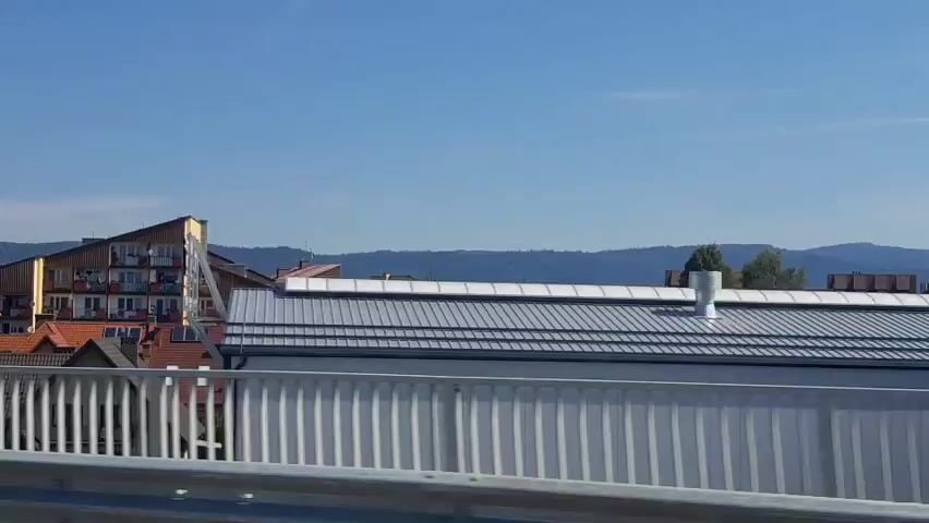 The video shows a view from a bridge with a metal railing in the foreground. In the background, there are several buildings, including a large, modern, multi-story building with a slanted, wooden facade and balconies. There are also houses with red tile roofs and some trees. The sky is clear and blue, and there are hills or mountains in the distance. The video appears to be taken from a moving vehicle, as indicated by the slight blurring and the changing perspective of the scene.