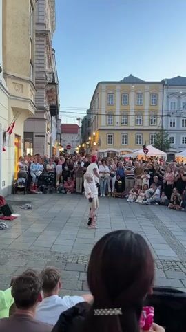 The video captures a vibrant street performance scene in Linz, Austria. The primary subjects are a crowd of people gathered to watch two performers in the foreground. The performers, one appearing older with white hair and wearing a white t-shirt and shorts, and another, possibly younger, in a red hat and darker clothing, are in the center of the cobblestone street, seemingly engaged in an act that draws the attention of the onlookers.

The scene is set in a European city street lined with multi-story buildings. To the left, a building with a yellow facade and dark trim is prominent, with a red and white flag hanging from its upper level. Across the street, a bright yellow building with white window frames stands out, and further down, a grey building with a similar architectural style is visible. The sky is a clear, pale blue, suggesting it is either early morning or late evening, with the lighting indicating dusk.

The crowd is diverse, comprising people of all ages, many of whom are sitting or standing to get a better view of the performance. Some are captured from behind, offering a perspective of being part of the audience. The atmosphere appears lively and engaged, with the audience captivated by the performance. There are no explicit emotional expressions easily discernible from the distance and image quality, but the gathering itself suggests interest and enjoyment.

Objects visible include strollers, suggesting families with young children are present. A white tent structure is visible in the background on the right side of the street, possibly part of a market or event. There is some faint text on signs on the buildings, but it is not legible in the provided frames. The overall impression is a public square or street that has been temporarily transformed into a venue for street entertainment, with the architecture and clear sky providing a picturesque backdrop.