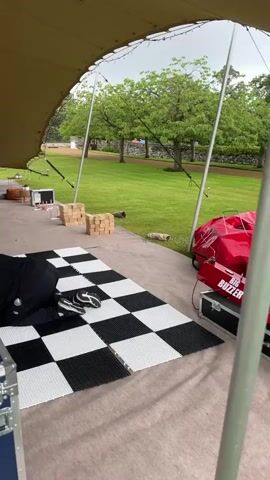 The video shows a large outdoor tent with a checkered dance floor laid out. A person in black clothing is on their hands and knees on the checkered floor. There are speakers and DJ equipment visible in the background, suggesting a setup for an event. Beyond the tent, a grassy area with trees and a stone wall is visible. There are also various seating options, beanbags, and games like cornhole set up, indicating a relaxed outdoor gathering or party. Another person is seen near the DJ booth in the background. A sign that reads "GARDEN GAMES SUMMER HOUSE" is also present.