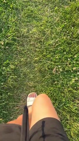 A first-person perspective video shows a person walking across a grassy area. The shot focuses on the lower half of the person's body, with their legs and feet visible. The person is wearing black shorts and white slide sandals. A dark-colored object, possibly a straw or a handle, is visible in the lower left corner of the frame.

The ground is covered in green grass, with some patches of clover and small white flowers. The lighting suggests it is late afternoon or early evening, with the sun casting a warm glow on the grass and the person's legs. The overall mood is casual and relaxed, as the person walks through what appears to be a park or a field. There is no visible text in the video. The location context is Zhodzina, Belarus.