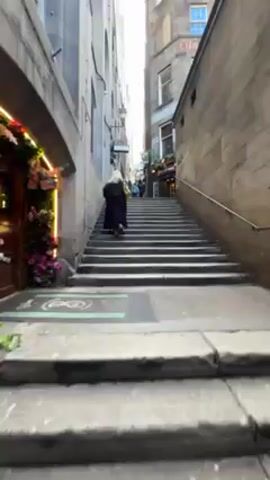 The video shows a narrow stone staircase in an urban environment. On the left is a building decorated with flowers and lights around its entrance. A person in a long black coat is walking up the stairs. Other people are visible further up the staircase. Buildings surround the staircase, one displaying the word "SUBURB" on its facade. A pub named "HALFWAY HOUSE" is visible halfway up the stairs. The day is overcast.