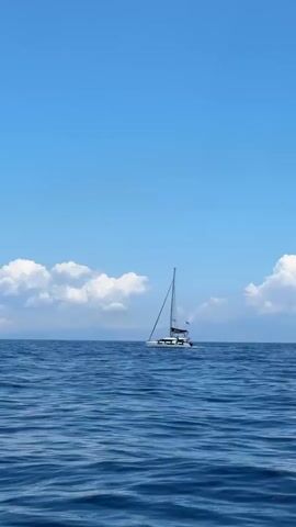 The video captures a serene daytime scene on the open sea near Koundouros, Greece. A white catamaran sailboat, featuring a single mast and a small flag, is the primary subject, moving slowly across the calm, deep blue water. Its sails are not deployed, indicating it is likely motoring. The sky is a clear, vibrant blue, adorned with fluffy white cumulus clouds stretching across the distant horizon, beneath which hazy land or mountains are faintly visible. The water surface displays gentle ripples, reflecting the bright sky. The camera, presumably aboard another vessel, subtly sways with the waves, accompanied by the peaceful sound of water against a hull. No people are visible, and there is no discernible text.