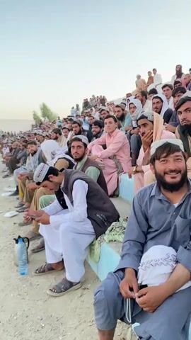 The video shows a crowd of people, predominantly men, sitting on bleachers outdoors. They are dressed in traditional clothing, including shalwar kameez and kufi hats. The atmosphere appears casual, with some individuals looking at their phones while others engage in conversation. The lighting suggests it is daytime, possibly late afternoon. The ground is dusty and appears to be a sports field or stadium. A few water bottles are visible on the ground. The individuals in the foreground are looking towards the camera, with one man smiling. The crowd is densely packed, indicating a significant gathering.