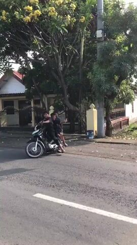 The video shows three young individuals riding a black Honda motorcycle side-by-side, with one person driving and the other two passengers seated behind. They are on a paved road in what appears to be a residential area, with houses and trees lining the street. The sunlight suggests it is daytime, possibly late afternoon given the shadows. The individuals are dressed casually in dark clothing. The driver is on the left, controlling the handlebars, while the two passengers are seated behind him. The middle passenger is looking at the camera and smiling, while the passenger on the far right is also looking towards the camera with a smile and their arms out. The overall mood appears to be lighthearted and fun. The video is short, capturing a brief moment of them riding together. There is some text visible on the side of the motorcycle, including the word "Honda". The setting appears to be a warm climate, typical of Kepatihan, Indonesia.