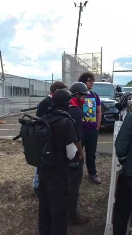 The video shows a group of people standing outside what appears to be a prison or detention facility.  Several of the individuals are wearing protective gear, including helmets, gas masks, and shields. Some individuals are wearing press badges. There are vehicles parked in the background.  The sky is overcast. The group appears to be engaged in a protest or demonstration.