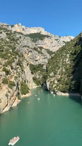 The video shows a scenic view of a turquoise lake surrounded by steep, rocky cliffs. The sky is clear and blue, indicating a sunny day. Several small boats and kayaks are scattered across the water, with some people visible on them. In the foreground, a white boat with several passengers is moving away from the camera. The cliffs are covered in green vegetation, suggesting a temperate climate. The overall impression is of a popular tourist destination offering outdoor recreational activities.