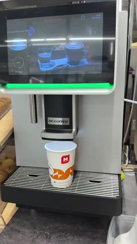 A close-up shot shows a hand holding a paper cup with an "M" logo and orange graphics under a coffee machine. The machine's spout dispenses a beverage into the cup, creating foam. The background shows the coffee machine's display screen and other objects.