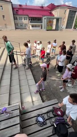A group of children and one adult are gathered on a set of outdoor concrete stairs. The adult, a man with light-colored hair wearing a green t-shirt and black shorts, is positioned on the stairs and holding onto two black resistance bands. Two boys, one wearing a white t-shirt and grey shorts, and another in a dark t-shirt, are also on the stairs, holding the other ends of the resistance bands and appearing to pull them. Other children are standing further up the stairs, observing. The environment appears to be a schoolyard, with a building visible in the background.