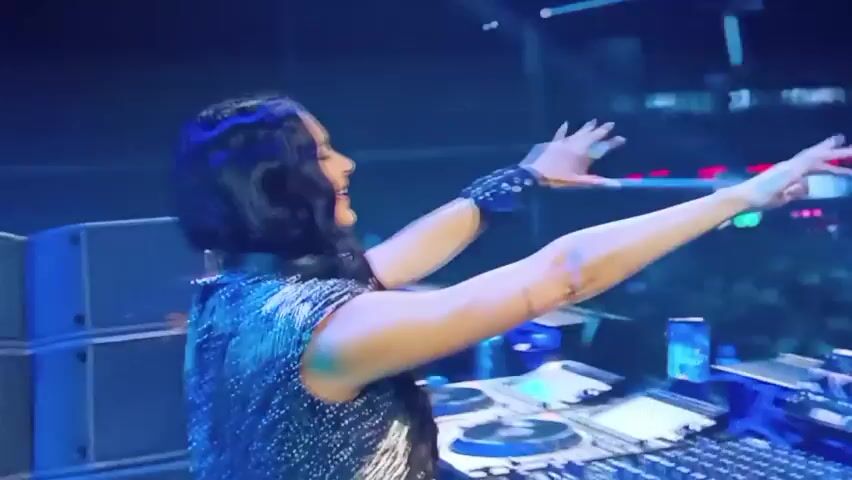 The video shows a dark, crowded environment with flashing lights. In the foreground, a person's face is partially visible, lit by blue and pink lights. Their eyes appear to be closed or looking down, and their hair is dark. Behind them, another person's blurry figure is seen. The background transitions to a wide shot of a concert or event with a stage, bright blue lights, and a large crowd. The atmosphere appears to be festive and energetic.