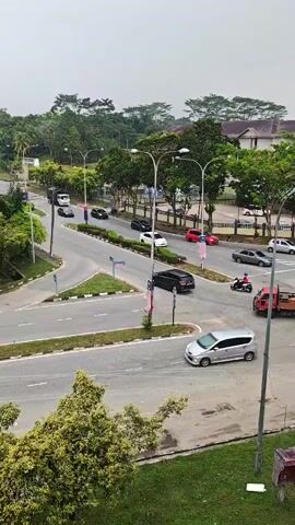 This is a drone-style shot looking down on a complex intersection with multiple lanes of traffic. It appears to be daytime, and the sky is overcast. The intersection is surrounded by lush green trees and some residential buildings. Several cars, including a red truck and a white van, are visible on the roads, navigating the intersection. There are also a few motorcycles. The road surfaces are asphalt, and there are clear lane markings and traffic signals. The overall scene depicts a busy urban or suburban intersection.