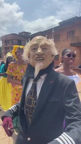 A person in a wrinkled, elderly mask with white hair and a white goatee stands in the foreground, wearing a black suit jacket, a black and gold tie, a black scarf, and maroon gloves. Behind them, a group of young women in colorful attire appear to be participating in a parade or celebration. One woman in the background wears a bright yellow skirt and a floral top, with a yellow headpiece. Another woman to the left wears a turquoise top and a long yellow skirt, also with a headpiece. The environment appears to be an outdoor street scene with buildings visible in the background. The sky is cloudy.