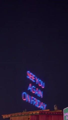 A sign with neon lights spelling out "SEE YOU AGAIN ON FRIDAY" is visible against a dark night sky. The letters are a mix of blue and pink. The sign is positioned above a red structure, possibly a building or a stall. To the right of the sign, a green sign with white lettering is partially visible. The overall scene appears to be outdoors, likely at an event or venue.