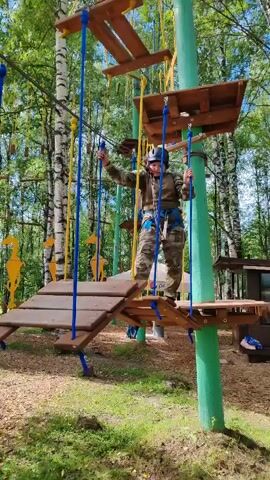 A person wearing camouflage clothing and a helmet is navigating an obstacle course in a wooded area. They are on a wooden platform suspended by ropes and are holding onto ropes for balance. The obstacle course appears to be made of wood and rope, with several platforms and suspended elements. The background shows trees and greenery, suggesting an outdoor park or adventure course. The person is moving from one platform to another.