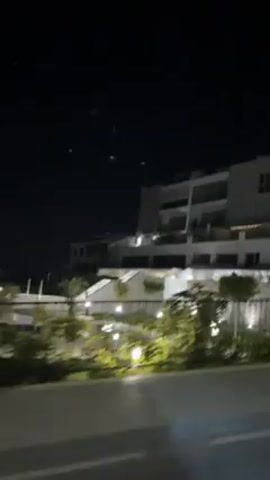 The video shows a person driving a vehicle, possibly a motorcycle, at night. Initially, a modern building with multiple stories and balconies is visible. Then the camera focuses on a black helmet with a colorful scarf or neck gaiter. Following this, the perspective switches to the driver's point of view, showing a road illuminated by street lights, with buildings visible in the distance. A metal fence lines the road. The overall scene suggests a nighttime ride through a residential or resort area.