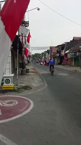The video shows a street scene in what appears to be a village or small town. The road is paved and lined with buildings on both sides. Red and white flags, possibly national flags, are prominently displayed, suggesting a celebration or event. A person is riding a bicycle in the foreground, heading away from the camera. Further down the road, several motorcycles and a red three-wheeled vehicle are visible, also moving away. The sky is overcast. The buildings appear to be a mix of shops and residences, some with peeling paint and signs. The overall impression is one of a normal day with some local traffic and decorations.