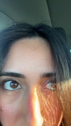 A close-up video shows a person's face from the forehead down to just below the nose. Their eyes are open and looking in different directions. The person has dark hair and brown eyes. Sunlight casts colorful shadows across their face. The background is blurred, suggesting the person is inside a vehicle.