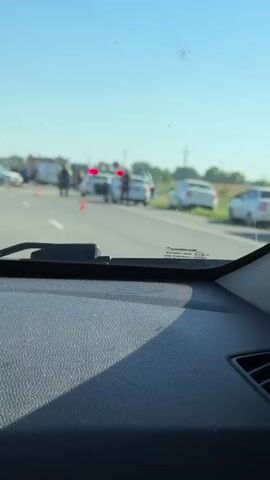 The video shows a blurry roadside scene from inside a vehicle. Traffic cones and emergency vehicles, including what appears to be a fire truck and police cars with flashing lights, are visible on the right side of a road. Several individuals, likely first responders, are standing near the vehicles. The sky is clear and blue, suggesting daytime. The overall impression is of a traffic incident or accident scene.